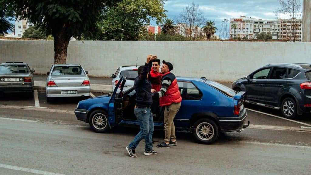 A group of three young men are gathered around a parked blue car, talking and gesturing.
