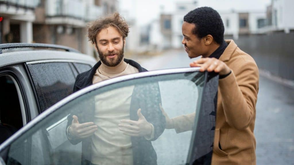 Two men stand by an open car door, one gesturing with his hands while the other smiles.