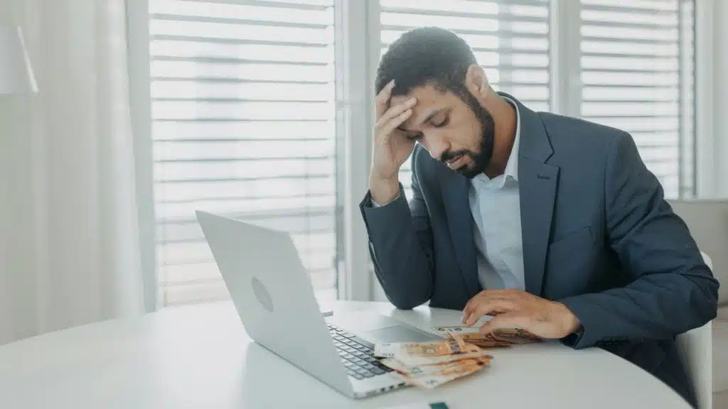 A sad man in a suit is sitting at a table with money, and a couple is on a sofa with their phones and a laptop.