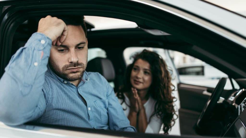 A worried man in a car, with a woman behind him with her hands clasped.