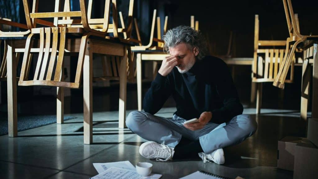 A man sits cross-legged on the floor, holding his head, surrounded by upturned chairs.