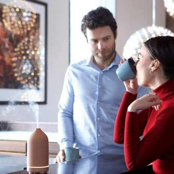A man watches a woman drink from a mug in a kitchen with a diffuser on the counter.