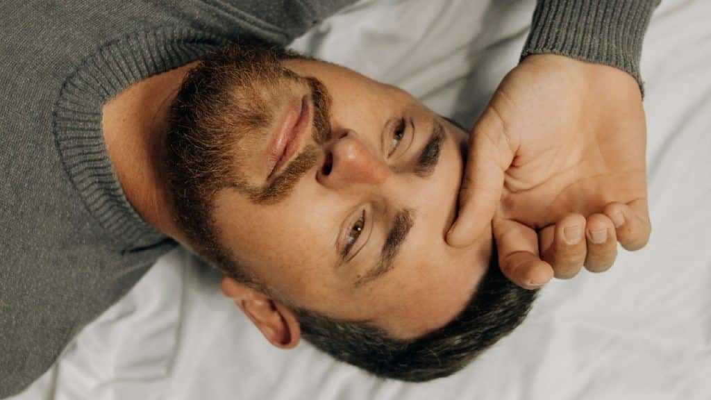 A bearded man in a gray sweater lies on a white surface, with one hand on his forehead.