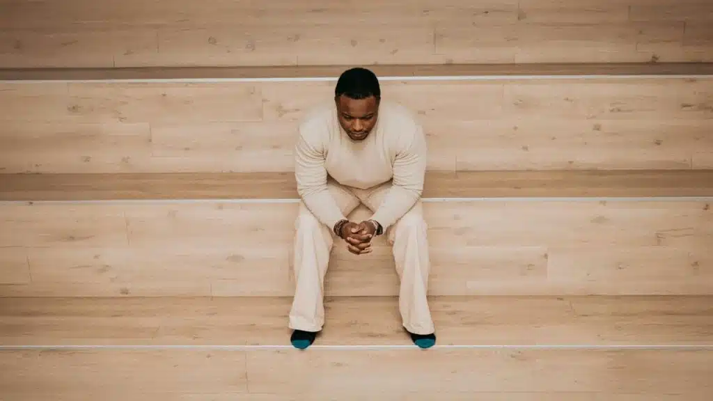 A man in a cream-colored outfit sits alone on a wooden bench, looking down with his hands clasped.