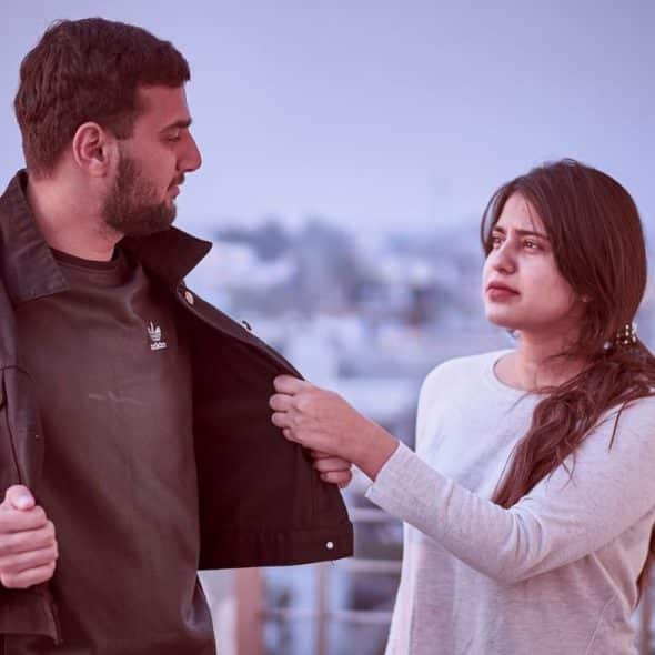 A man in a black jacket and a woman in a white shirt look at each other.