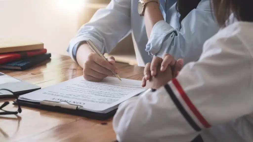 A person's hands point at a document on a table, with a pen and other hands nearby.