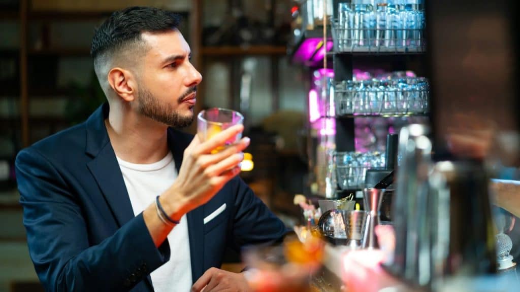 A man with a beard and dark hair sits at a bar, holding a glass.