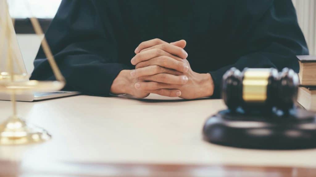 A person in black robes sits at a desk with their hands clasped, a gavel and scales of justice nearby.