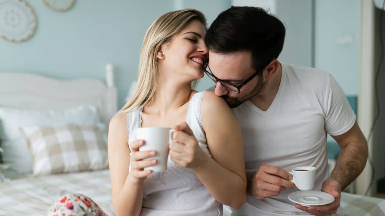 A man kisses a smiling woman's shoulder while they both hold cups.