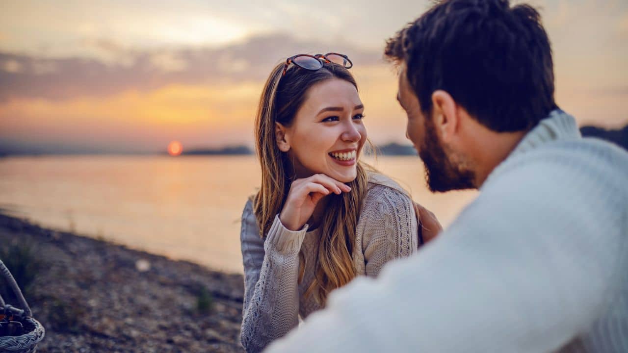 A smiling woman looks at a man by the water, with a sunset in the background.