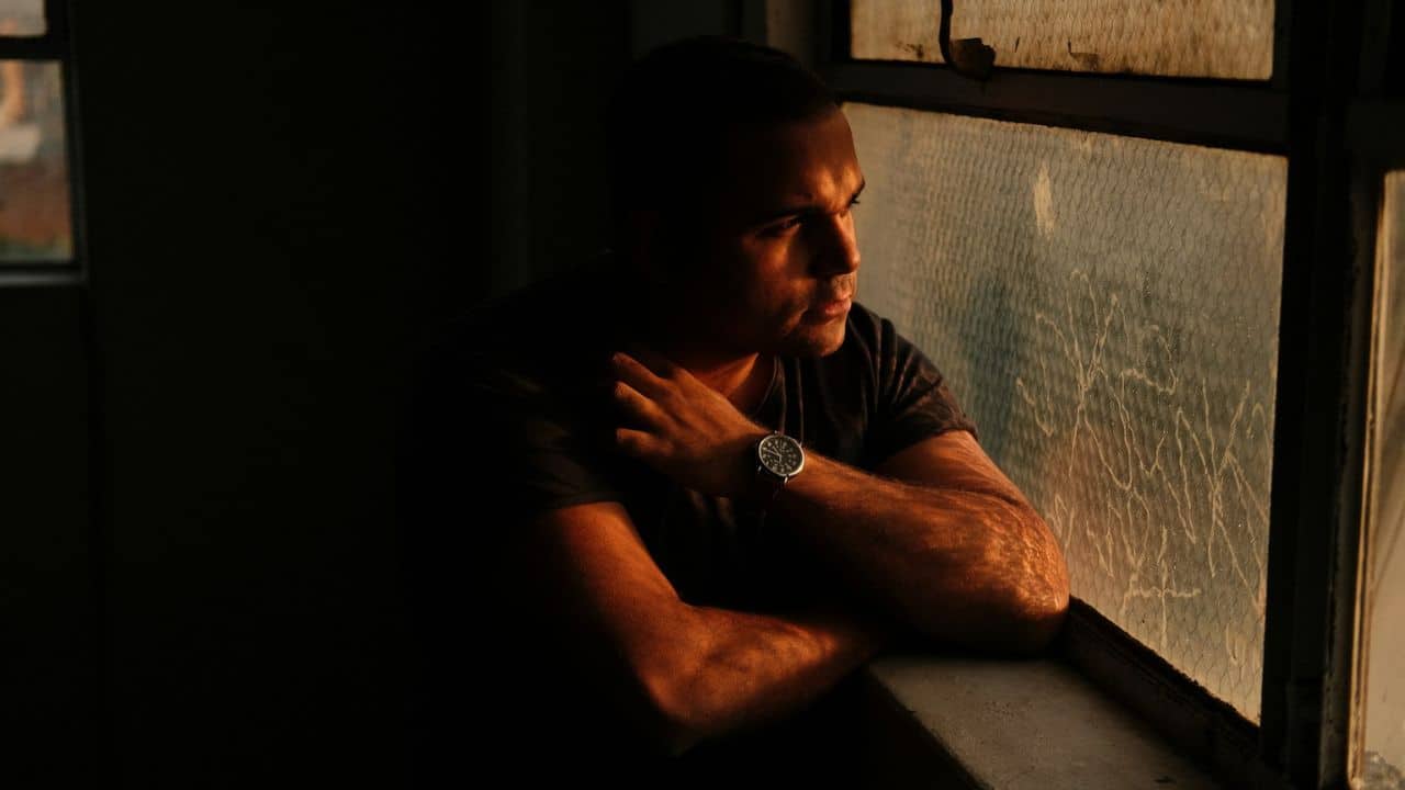 A man with short hair and a watch on his left wrist looks out a grimy window, illuminated by a warm light from the right.