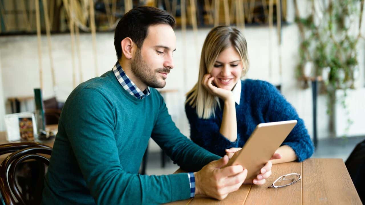 A man and woman sit at a table looking at a tablet together, with the woman smiling.