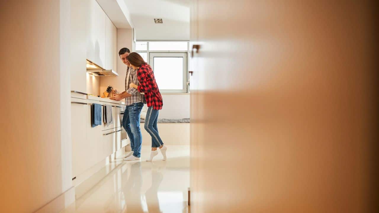 A man and woman are standing in a modern kitchen, with the woman leaning on the man.