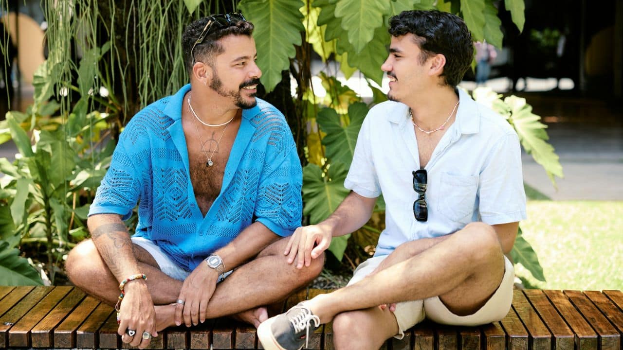 Two men are sitting cross-legged on a wooden bench, looking at each other and smiling.