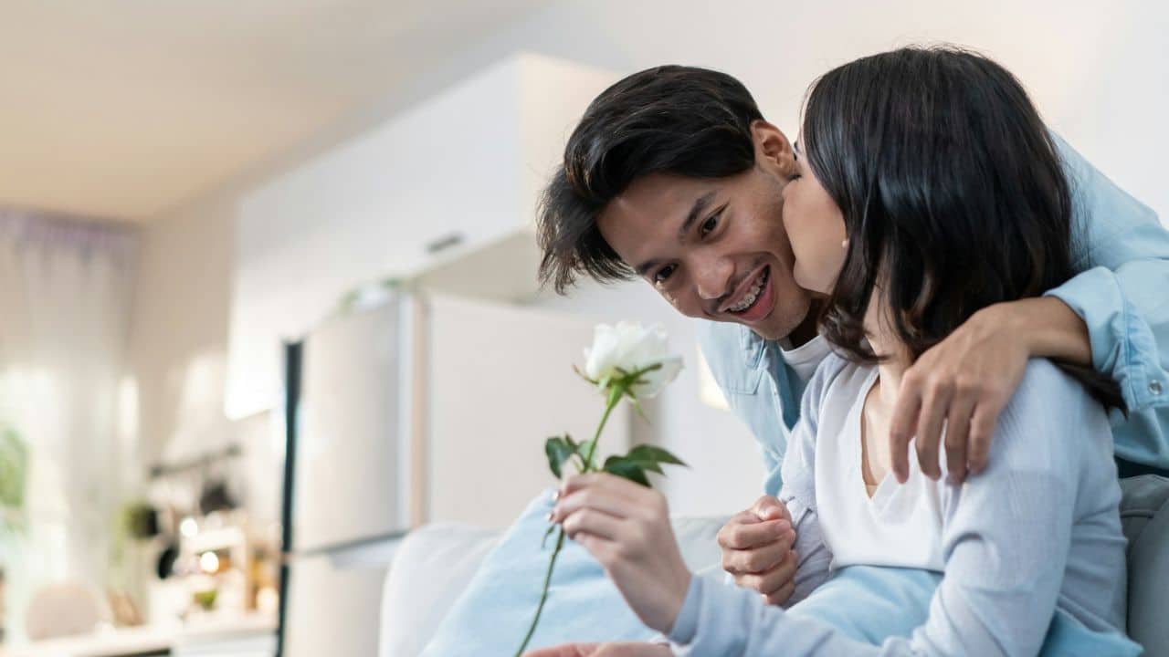 A man with braces leans in to kiss a woman on the cheek while she holds a white rose.