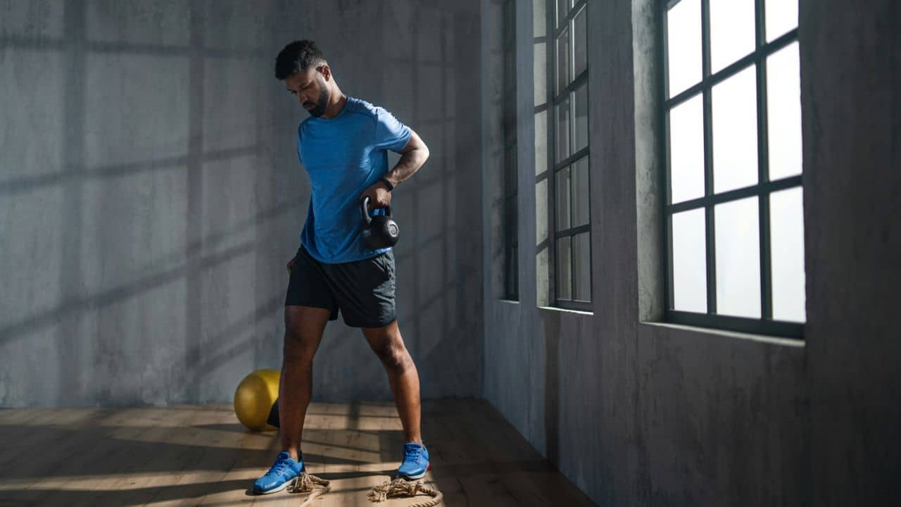 A man in athletic wear is holding a kettlebell in a gym with large windows.