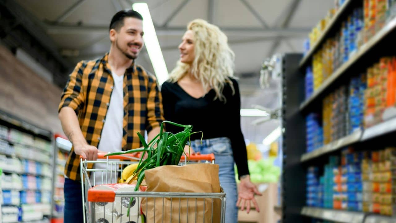 A man pushes a grocery cart while a woman with curly blonde hair smiles at him in a supermarket aisle.