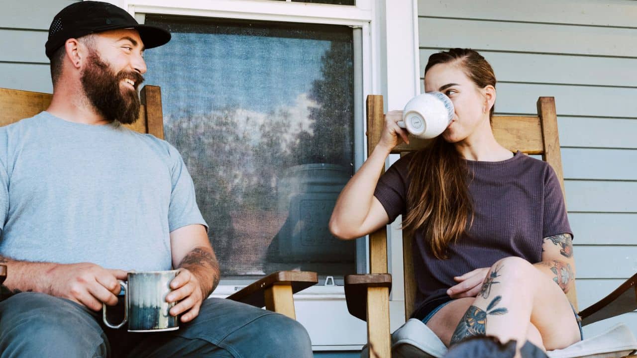 A man and a woman are sitting in rocking chairs on a porch, each holding a mug.