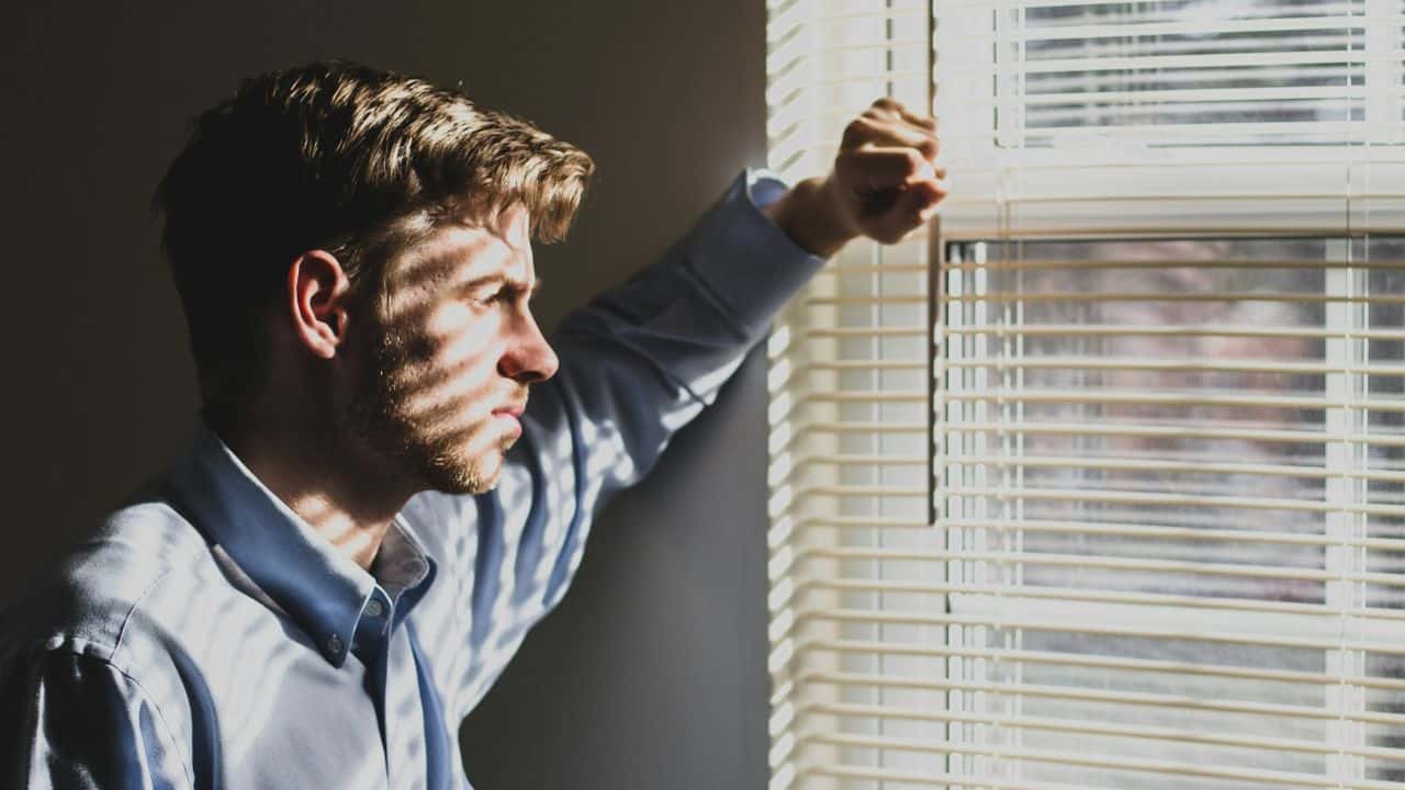 A man in a blue shirt looks out a window through blinds, light stripes on his face.