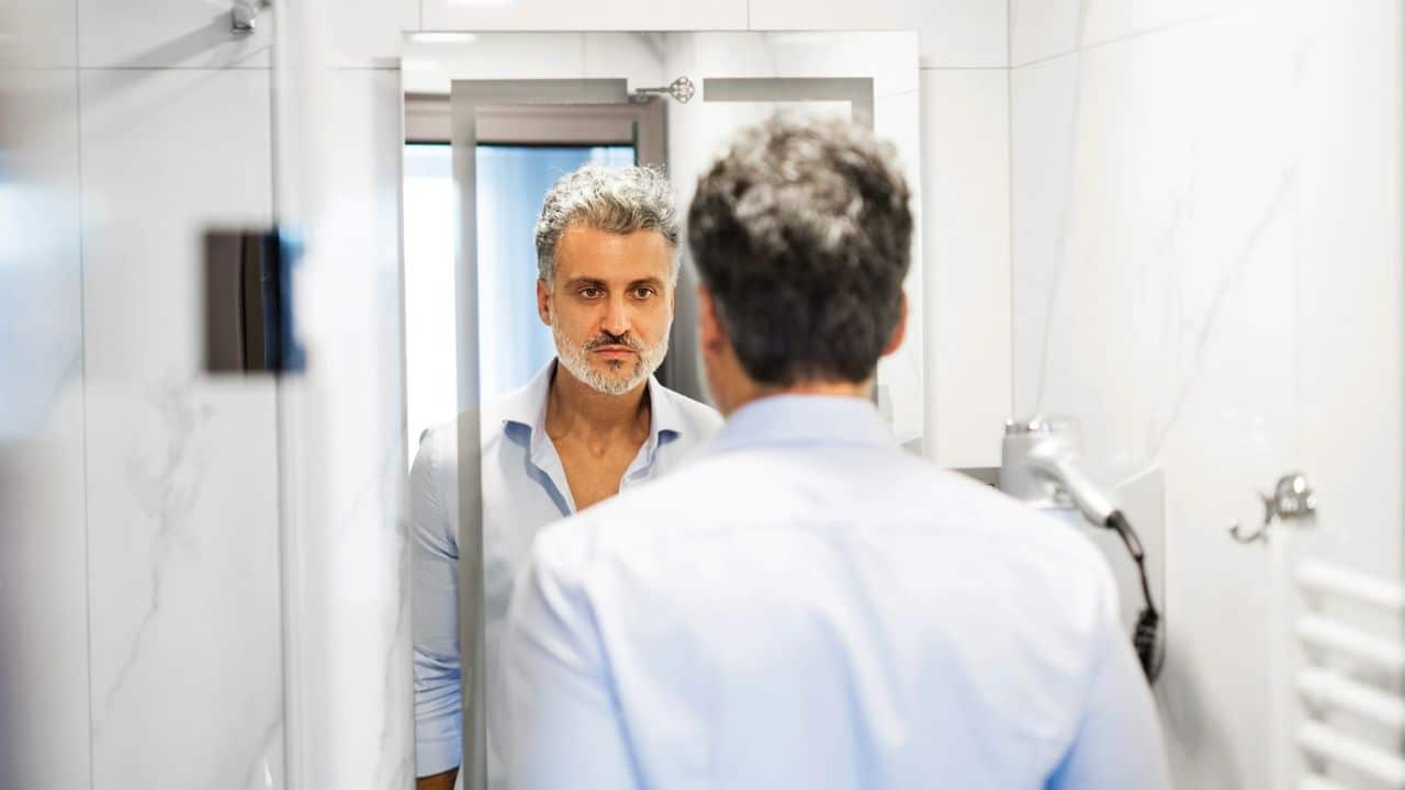 A man with salt and pepper hair looks at his reflection in a bathroom mirror.