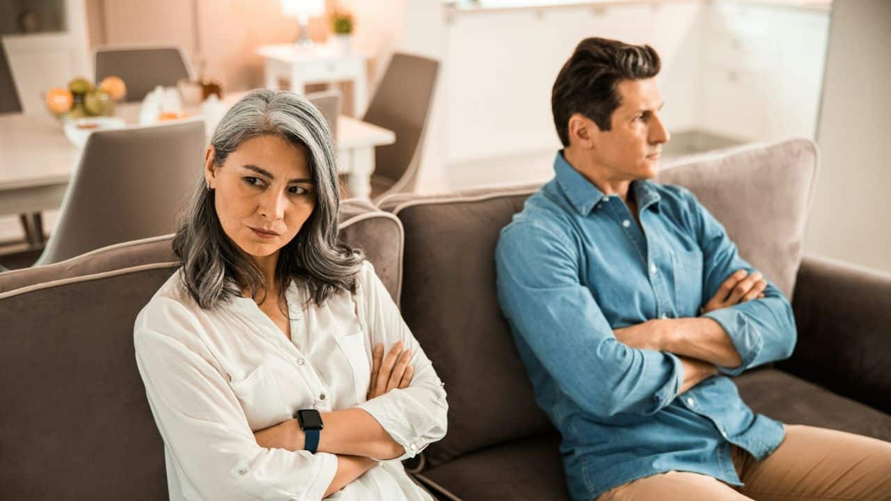 A displeased woman with folded arms sits on a couch, facing away from a man.