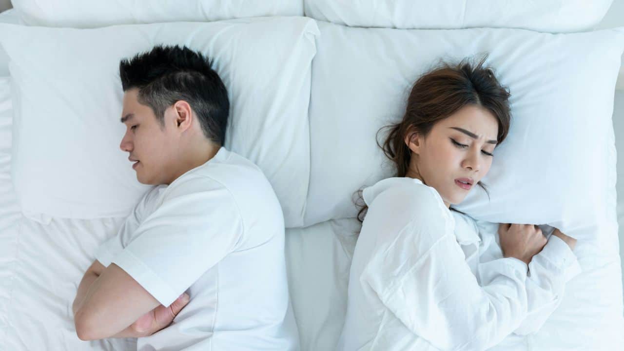 A man and a woman lie in bed facing away from each other, looking unhappy.
