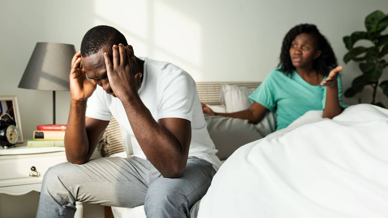 A man sits on a bed with his head in his hands, while a woman gestures angrily behind him.