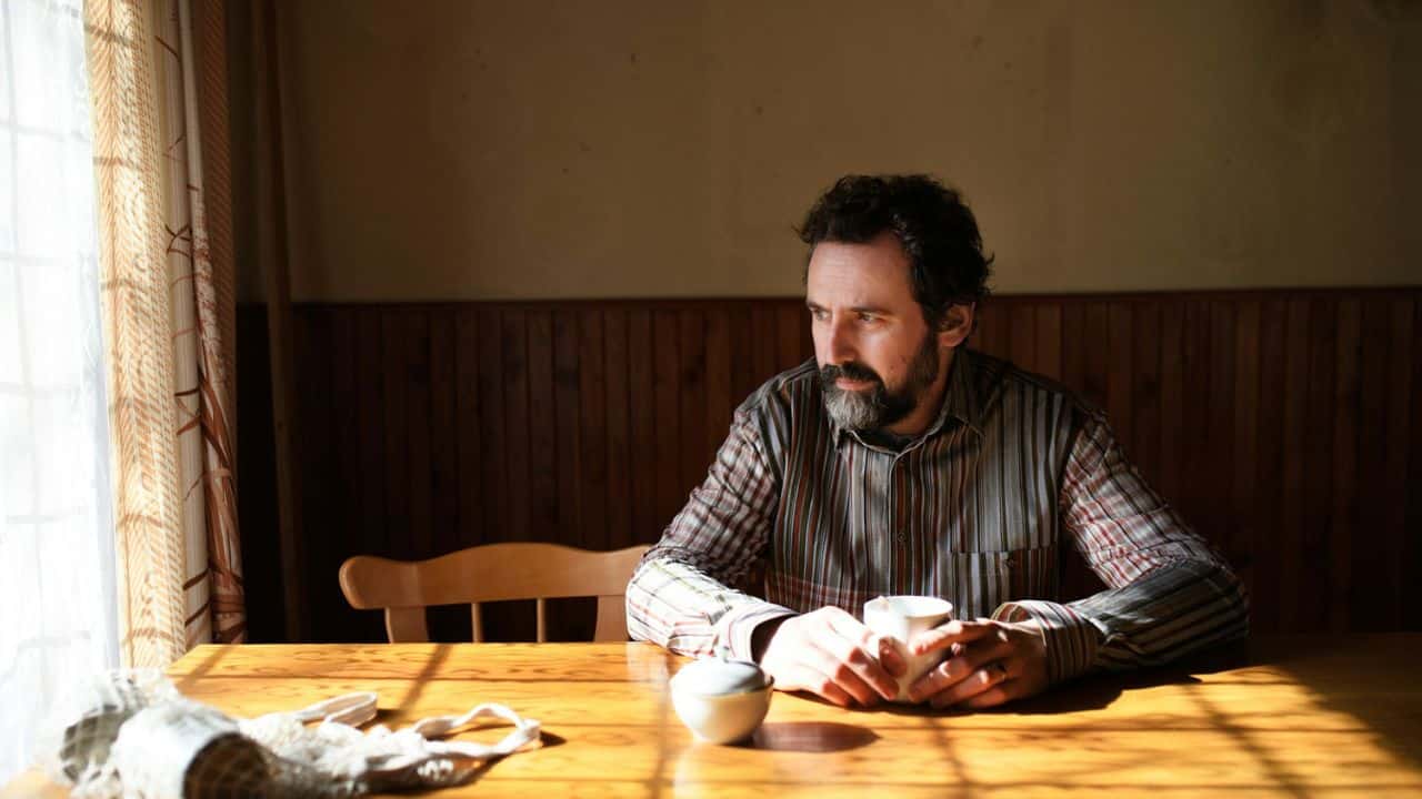 A man with a beard sits at a sunlit wooden table, holding a cup.