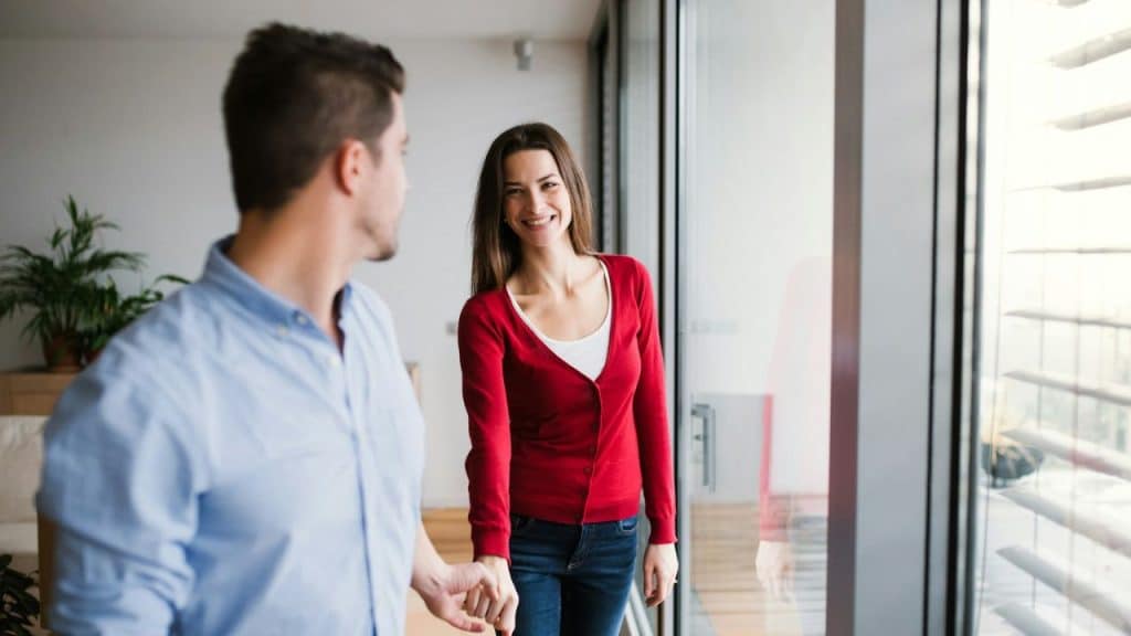 A couple holds hands and smiles at each other near a large window.
