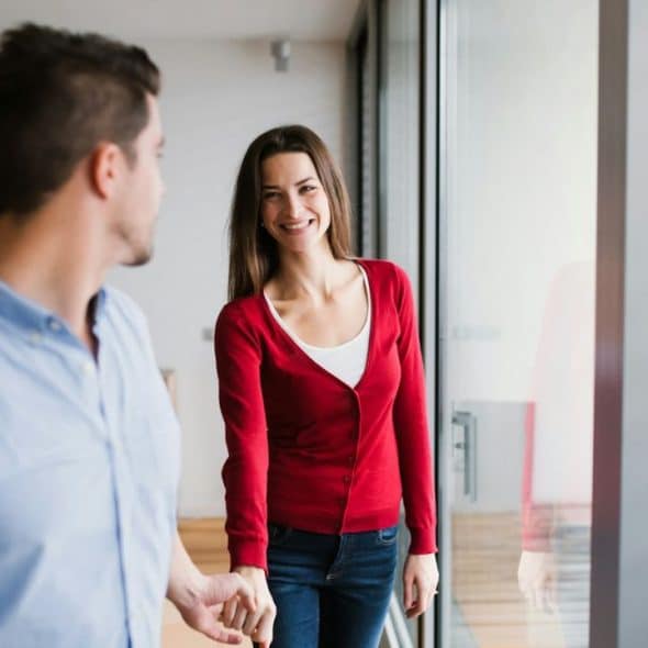 A couple holds hands and smiles at each other near a large window.