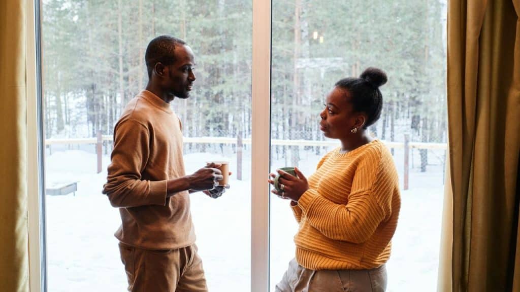 A couple stands near a window, holding mugs and talking to each other.