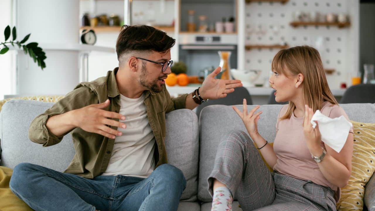 A man and woman sit on a couch, gesturing with their hands as they argue.