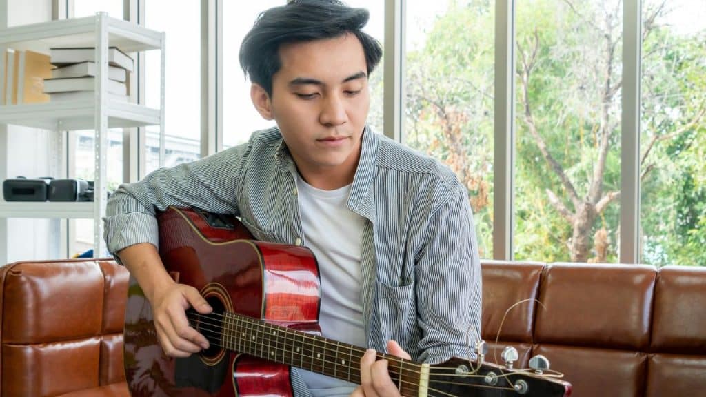 A young man with a striped shirt sits on a leather couch, playing an acoustic guitar.