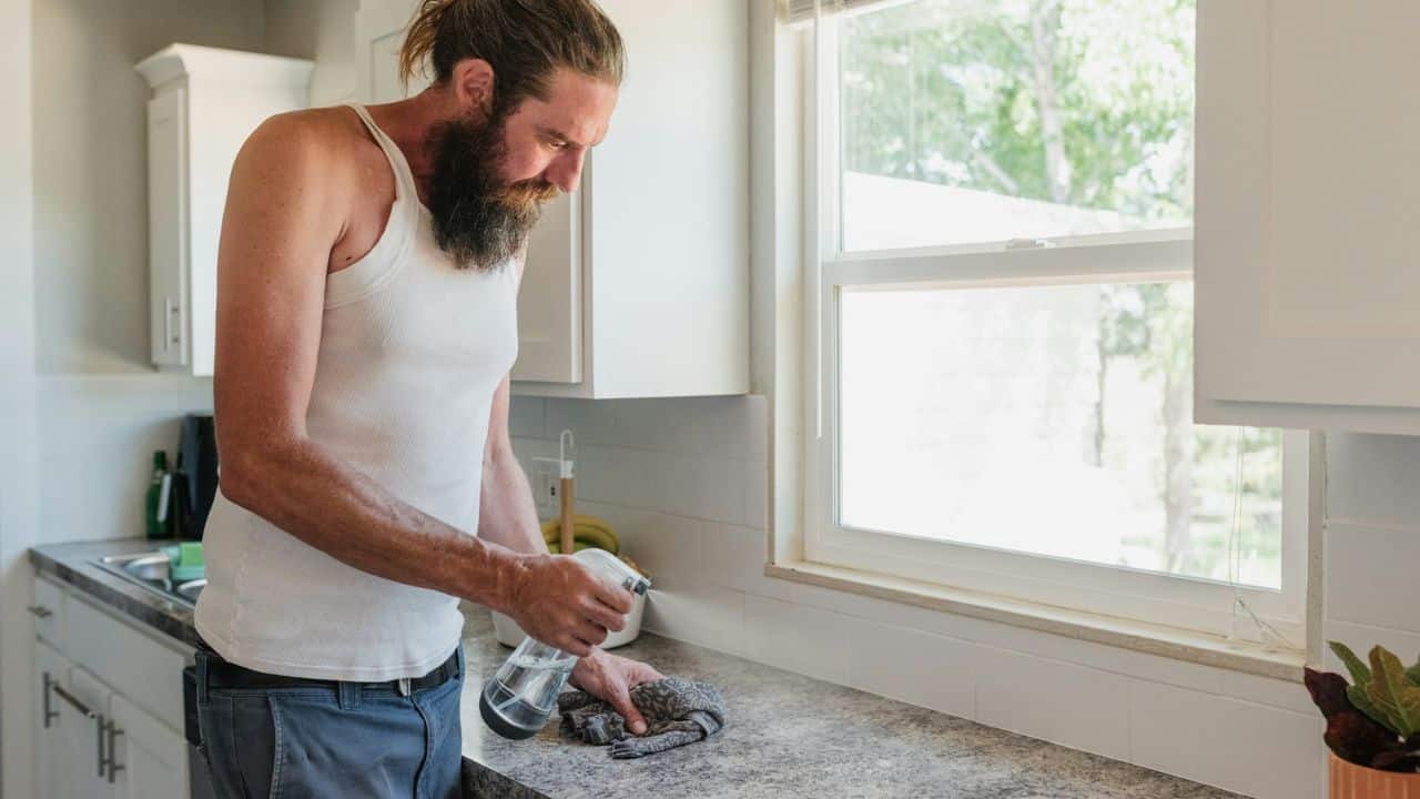 A man with a beard in a tank top cleans a kitchen counter with spray.