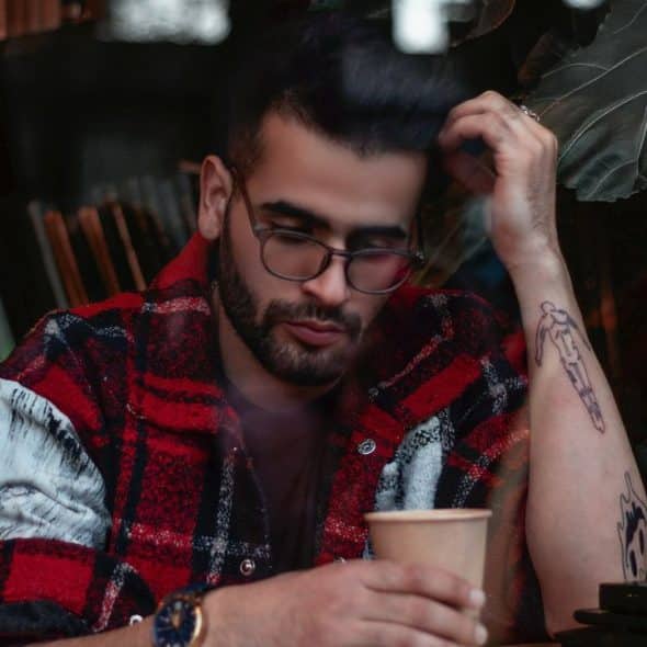 A man with a beard and glasses sits indoors, holding a paper cup.