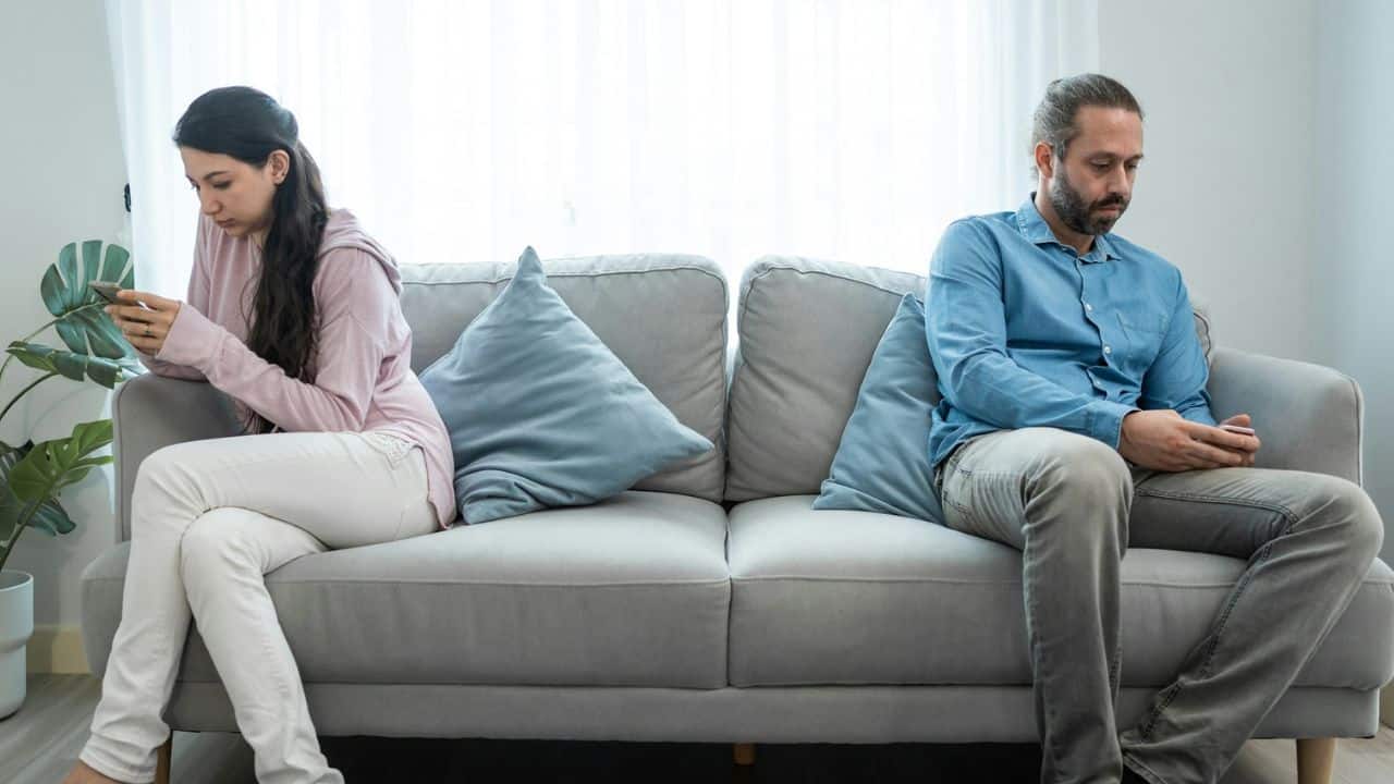 A man and woman sit separately on a couch, both looking at their phones.