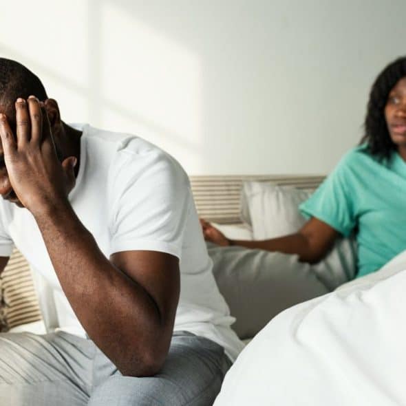 A stressed man holds his head in his hands while a woman argues with him in the background.