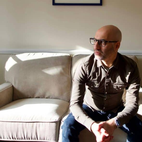A bald man wearing glasses sits alone on a couch, looking to the side.