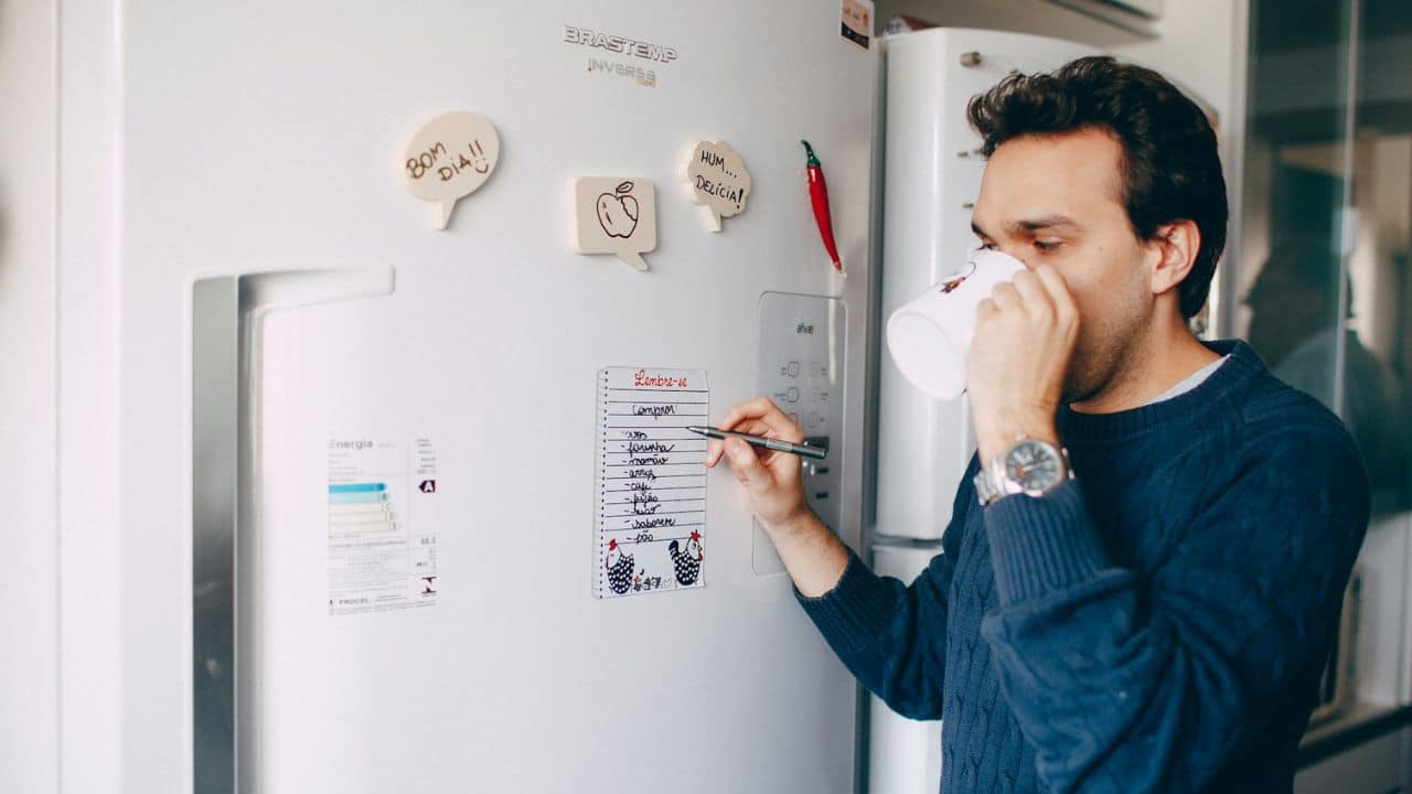 A man sips from a mug while writing on a to-do list magnet on a refrigerator.