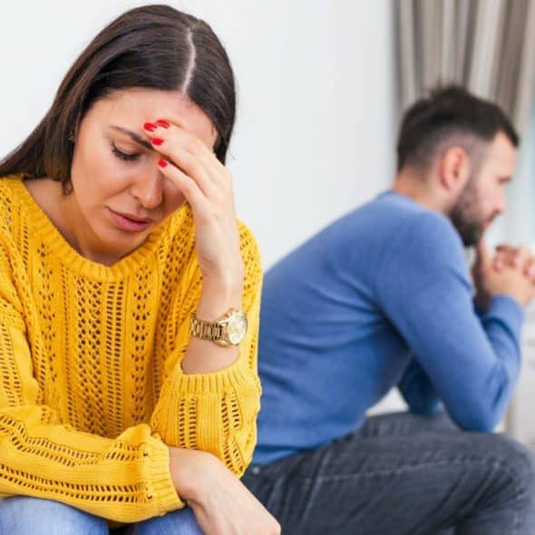 A woman in a yellow sweater sits on a couch with her hand on her head, looking away from a man.