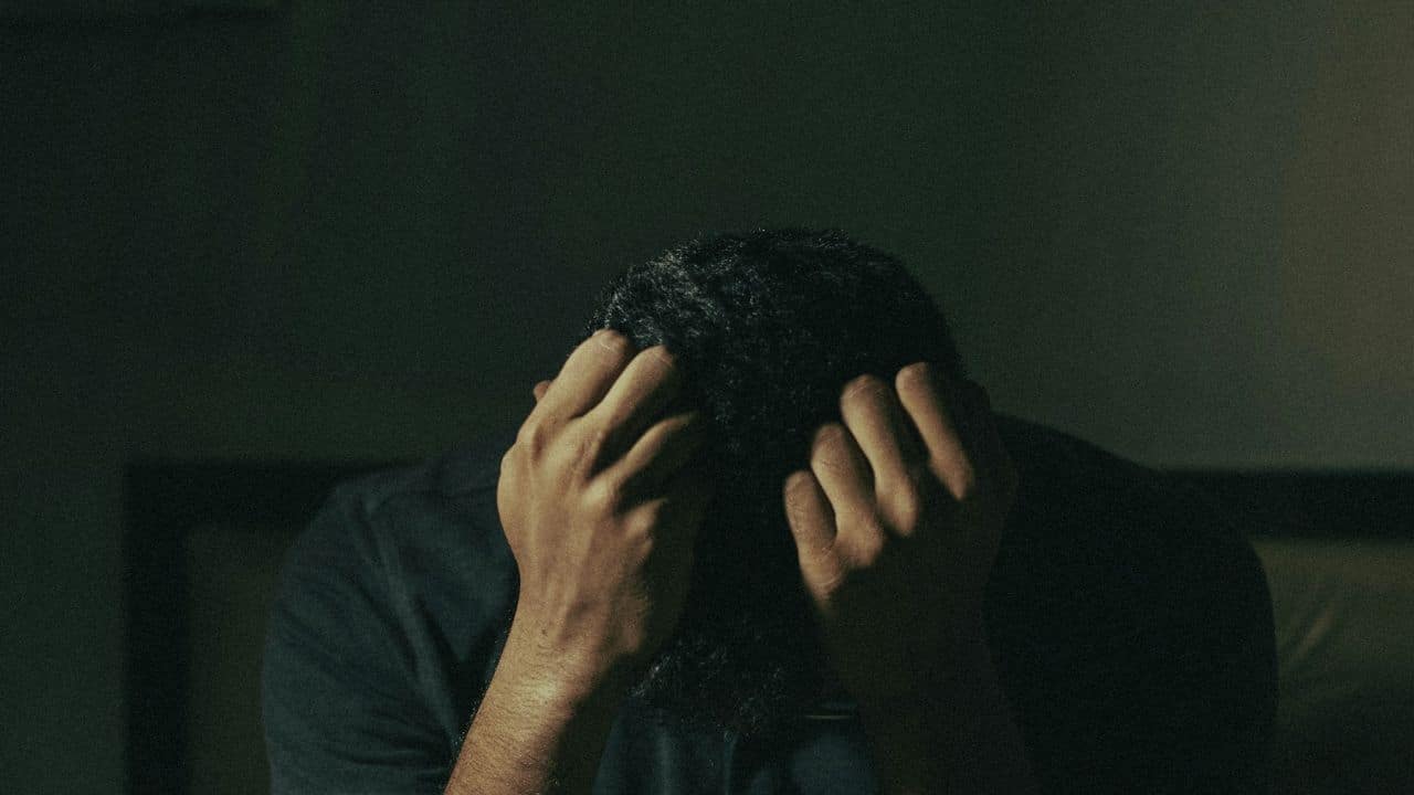 A person with dark, curly hair sits in a dimly lit room with hands covering their face.