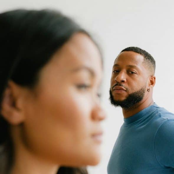 A blurry woman's face is in the foreground, with a man looking sternly in the background.