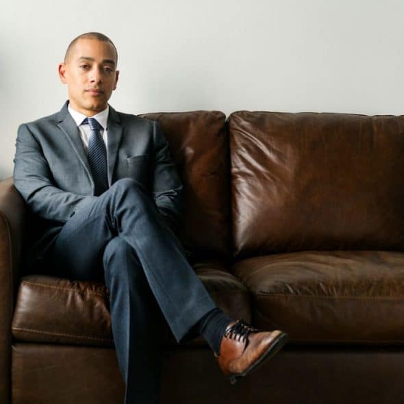 A man in a gray suit sits on a brown leather couch, looking at the camera.