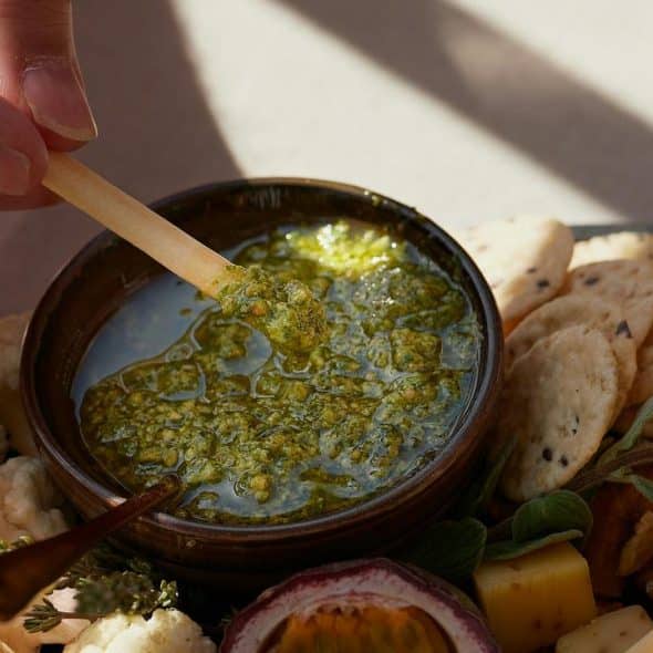 A person's hand dipping a cracker stick into a bowl of green pesto surrounded by snacks.