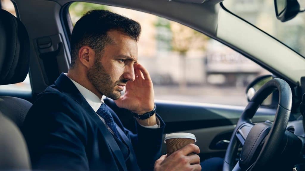 A man in a suit sitting in a car holding a coffee cup and touching his face.