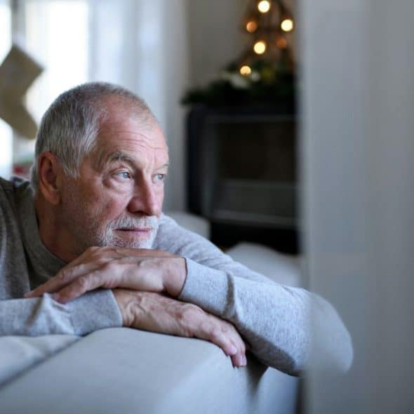 A lonely, elderly man with a beard sits on a couch, looking out a window.