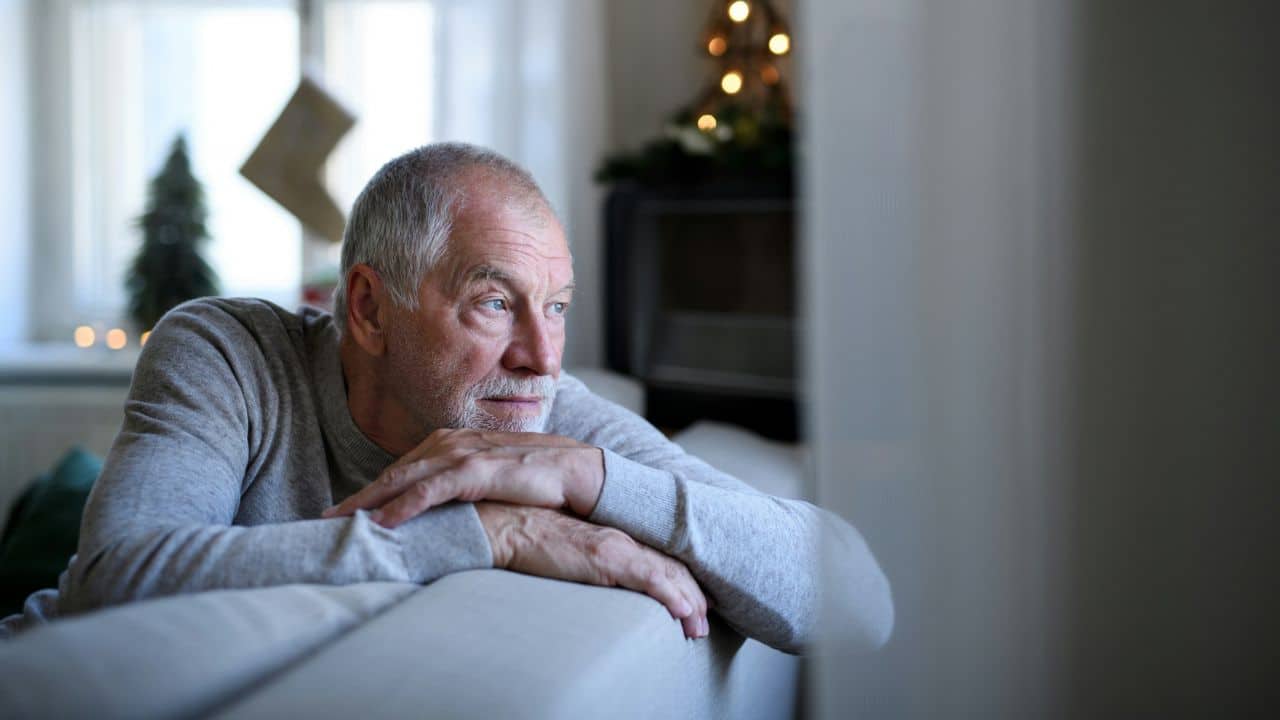 A lonely, elderly man with a beard sits on a couch, looking out a window.