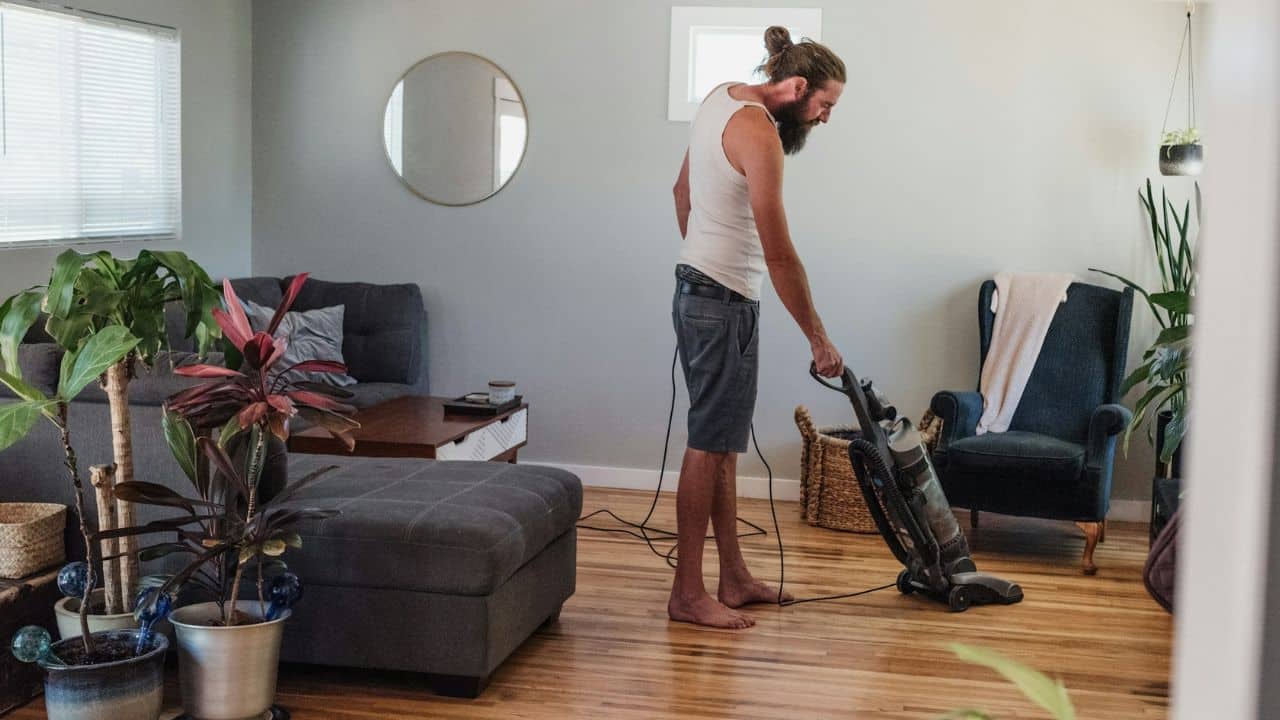 A man with a beard and hair in a bun vacuums the floor of a living room.