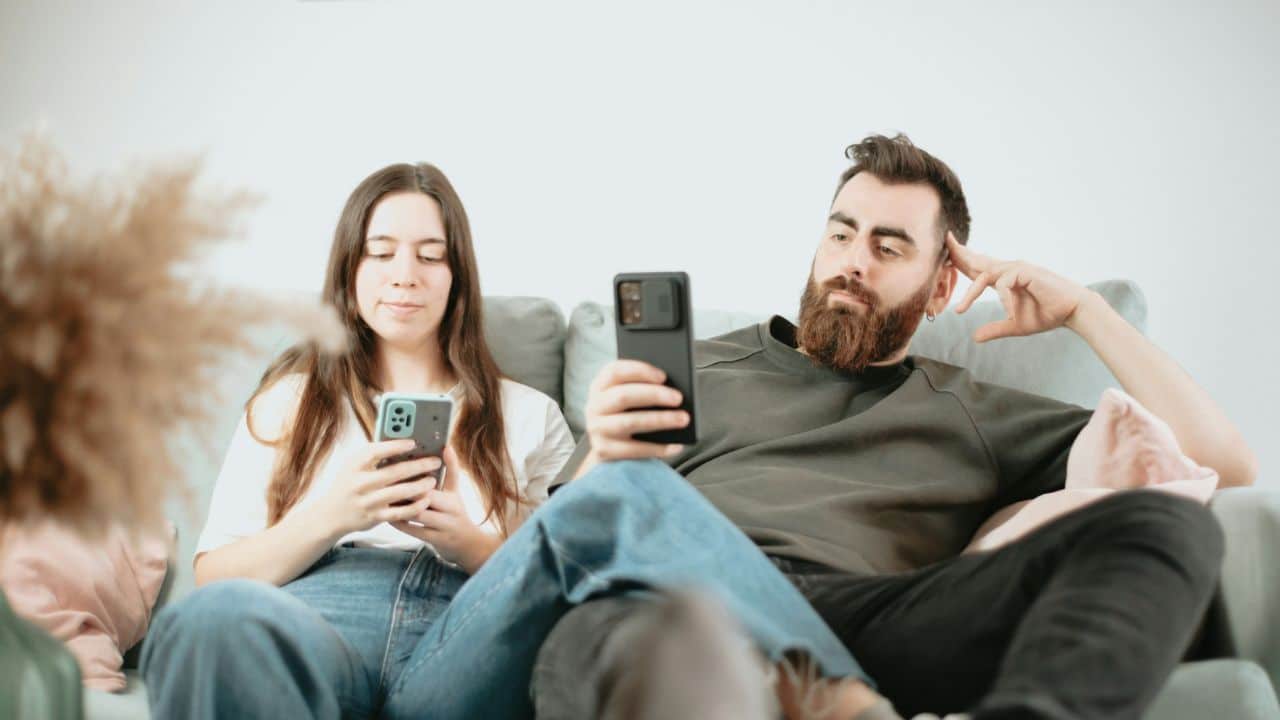 A man and a woman sit on a couch, both looking at their phones.