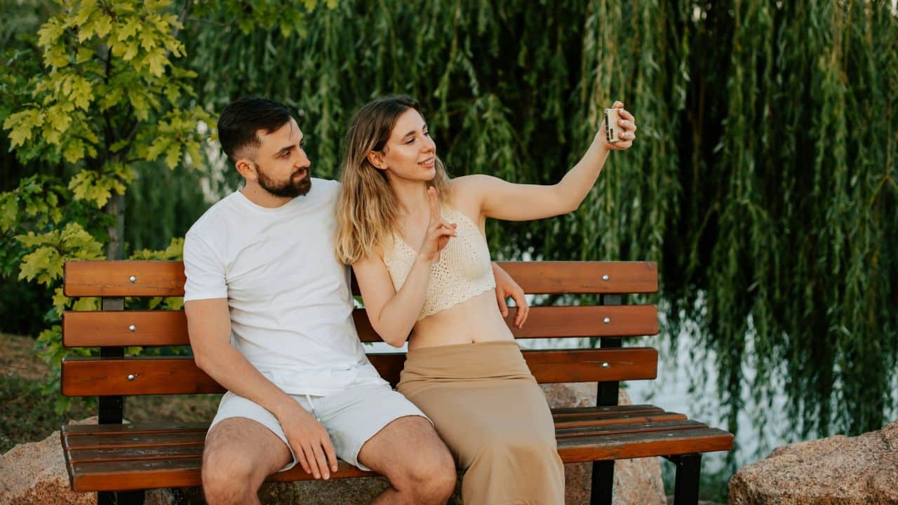 A woman takes a selfie with a man while sitting on a bench.
